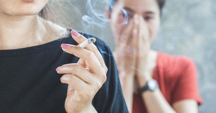Hand holding a lit cigarette while a person nearby covers their nose from the smoke